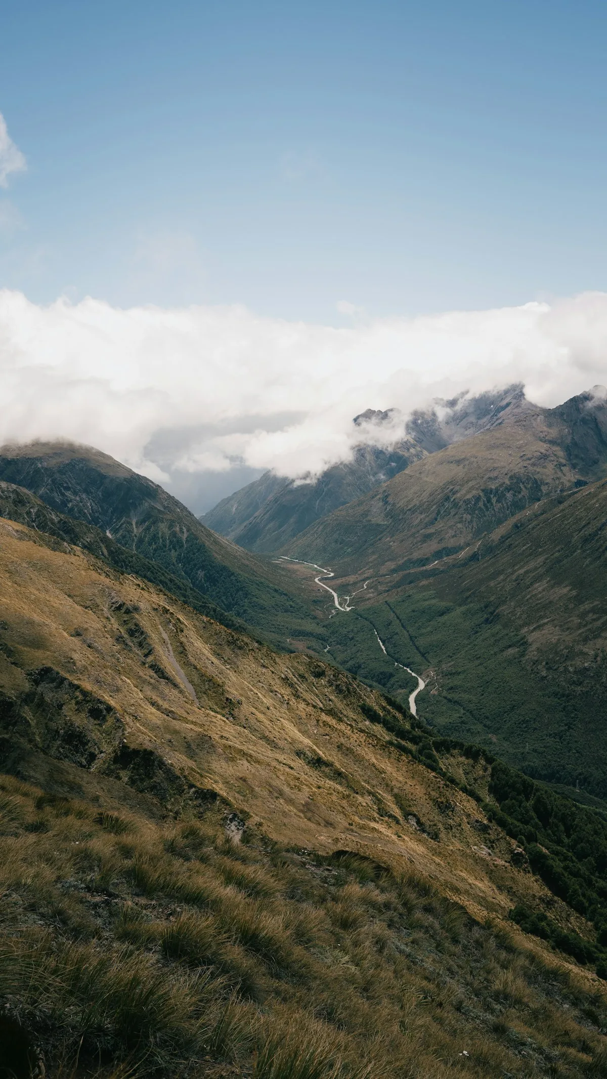 A winding pale river through a green and rocky mountain valley beneath low clouds and blue sky.